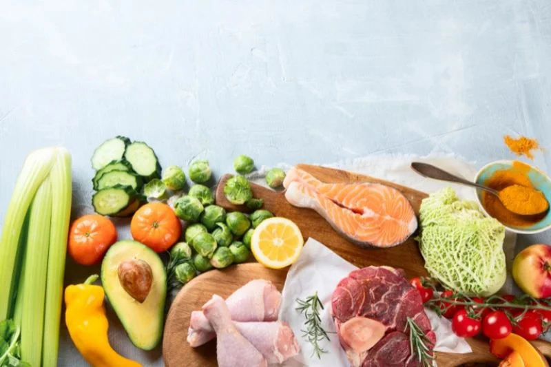Rau củ, cá hồi, thịt bò, thịt gà và trái cây tươi bày trên nền sáng cùng chén bột nghệ vàng