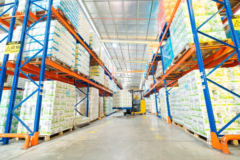 Lối đi giữa hai dãy kệ cao chất đầy pallet hàng trong kho của AIG, xe nâng chạy phía cuối