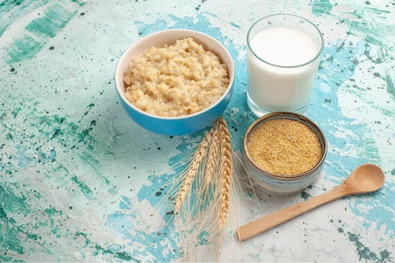 Bát cháo yến mạch, ly sữa và chén bột ngũ cốc mịn đặt cạnh bông lúa mì và thìa gỗ