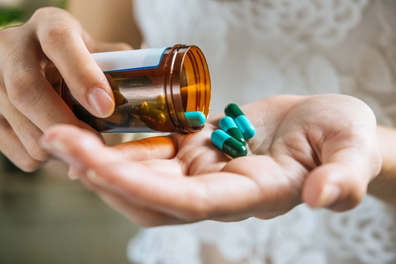 Bàn tay nghiêng lọ thuốc màu hổ phách, trút viên nang xanh và viên dầu cá vàng ra lòng bàn tay
