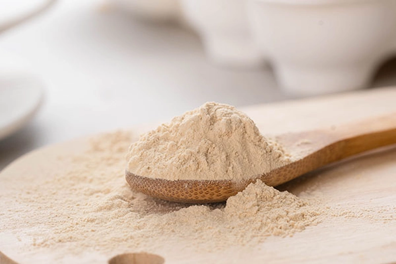 Thìa gỗ đầy bột Yeast Protein màu be đặt trên thớt gỗ, xung quanh là lớp bột rải mịn