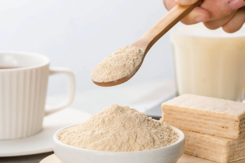 Tay cầm thìa gỗ xúc bột Yeast Protein bên bát sứ, cạnh tách trà và lát bánh xốp nhiều lớp